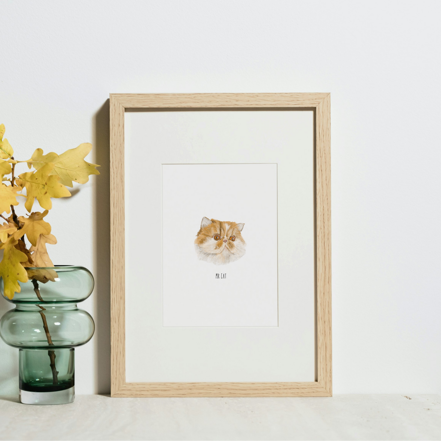 Framed artwork on a white surface with a vase of yellow leaves.