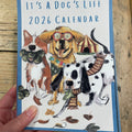 Calendar titled 'It's a Dog's Life 2026' held by a hand on a wooden surface