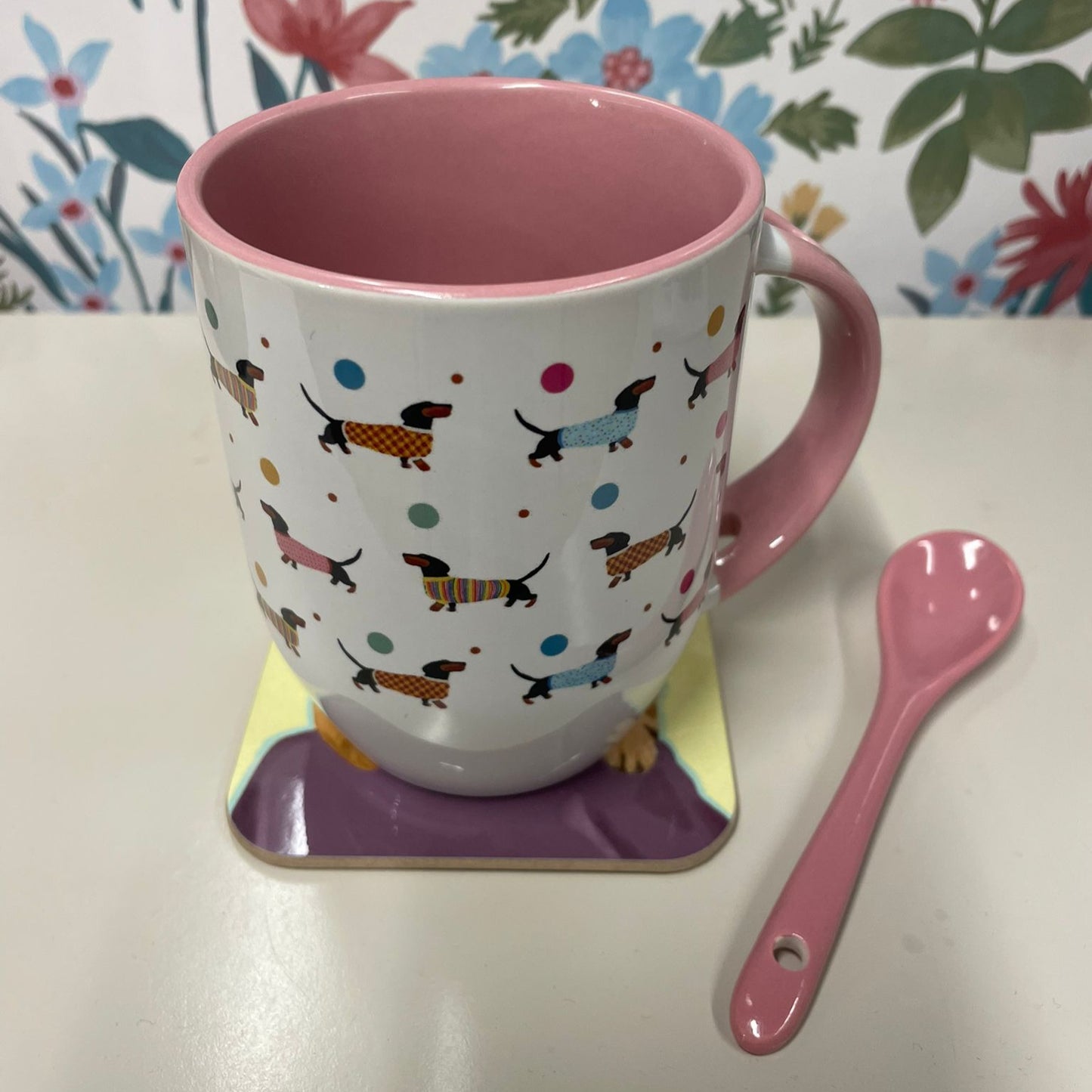 Colorful mug with dog pattern on a coaster, next to a pink spoon against a floral background