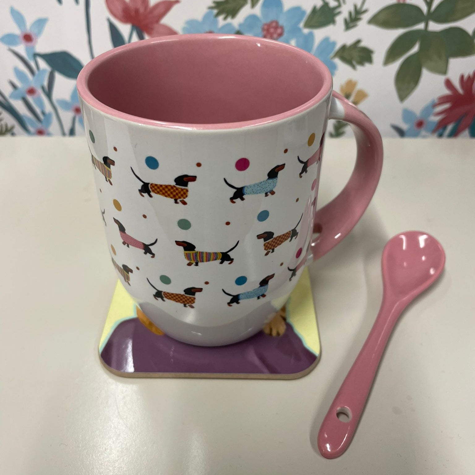 Colorful mug with dog pattern on a coaster, next to a pink spoon against a floral background