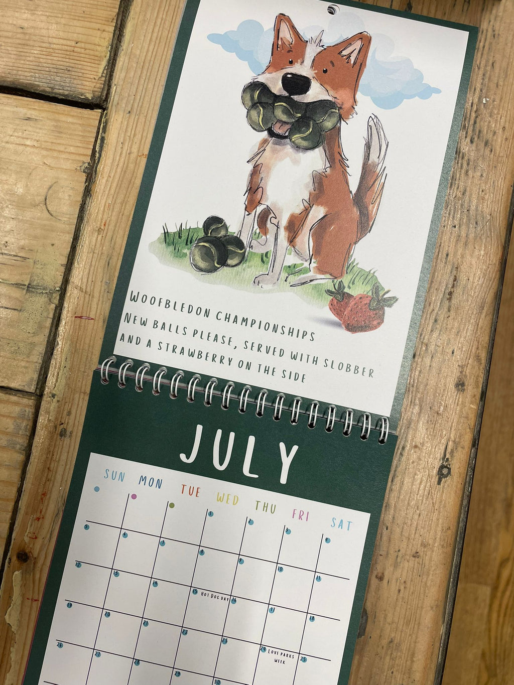 Calendar with July page showing an illustration of a Dog with tennis balls in his mouth  on a wooden surface