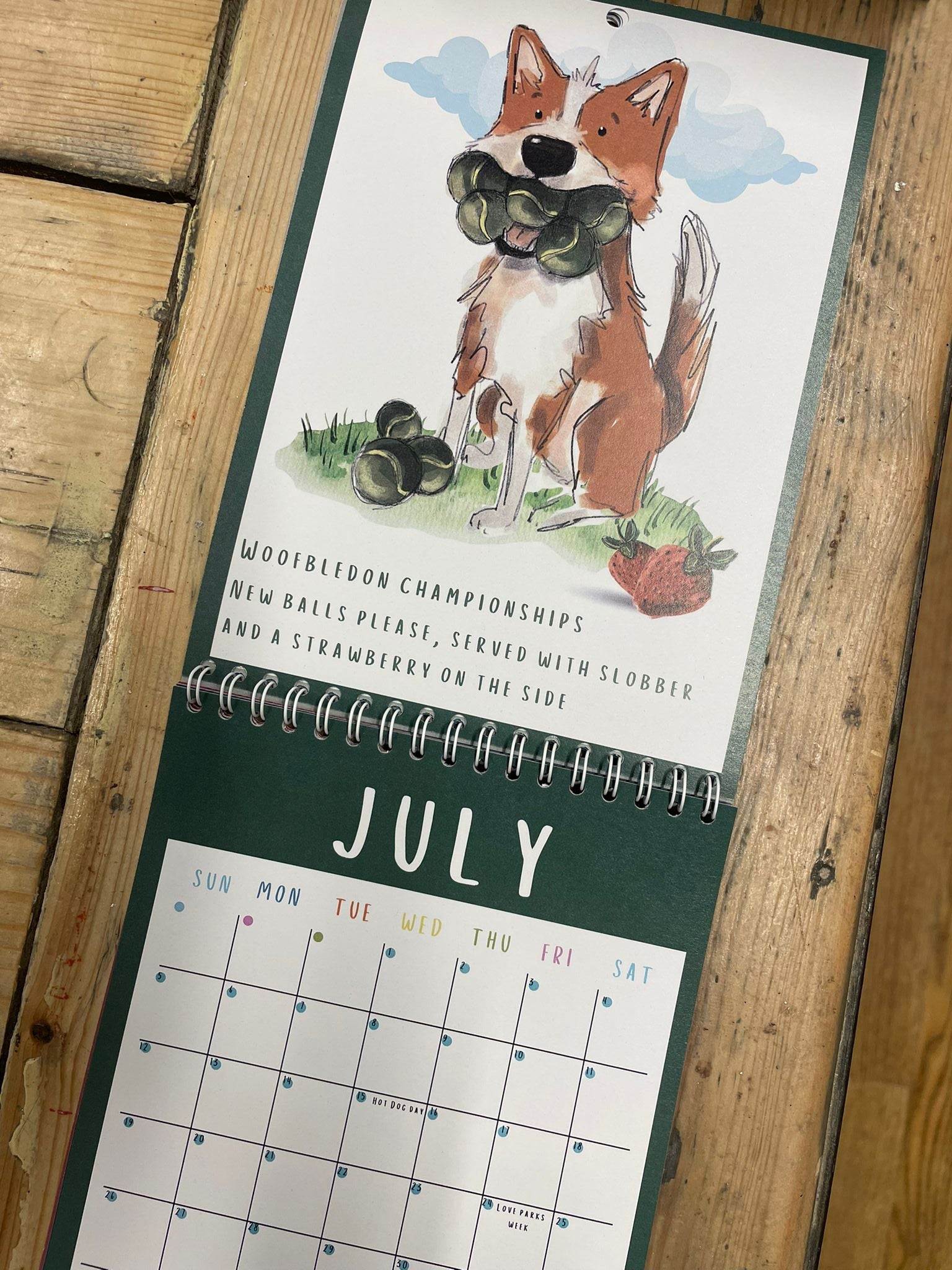 Calendar with July page showing an illustration of a Dog with tennis balls in his mouth  on a wooden surface
